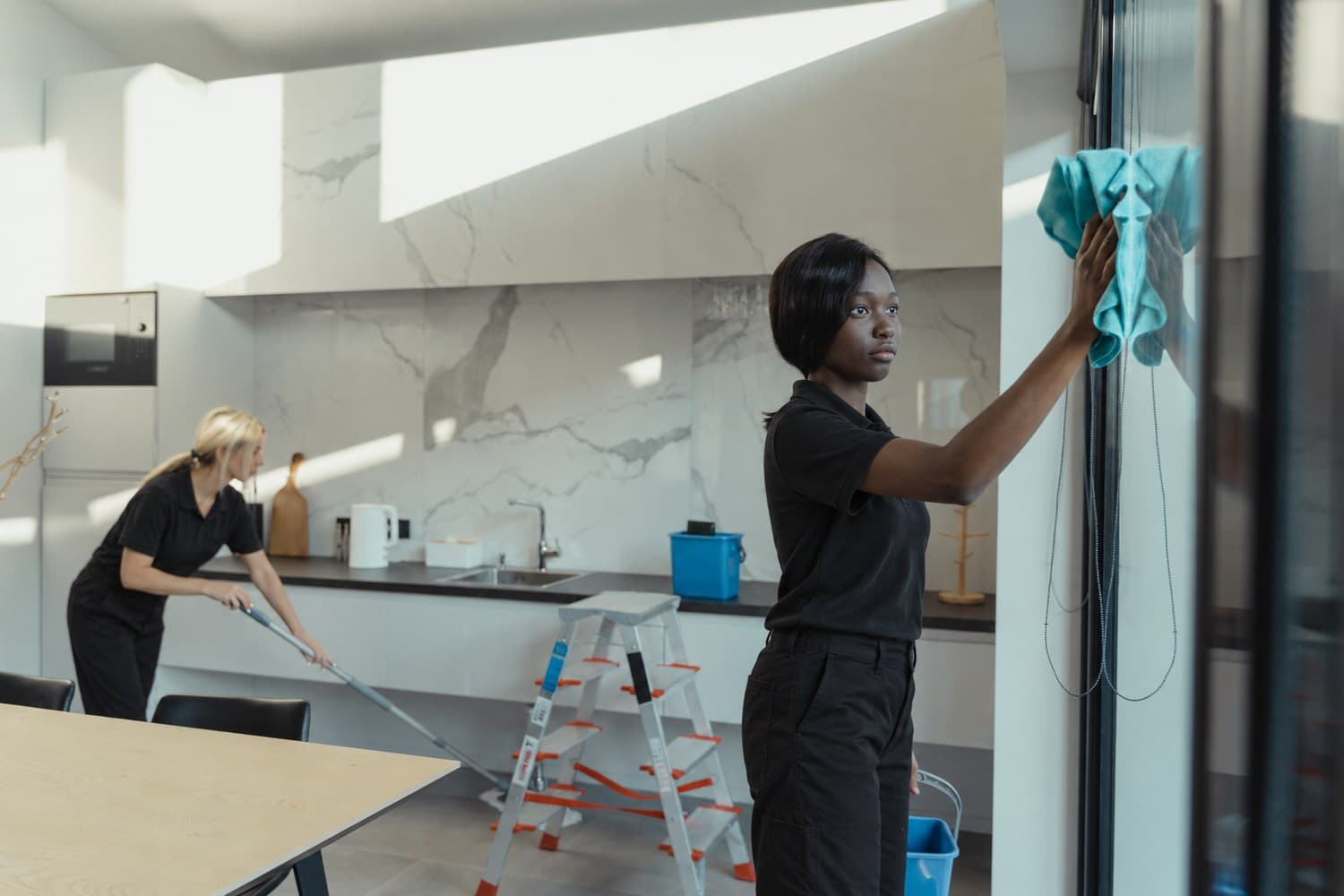 Two cleaners in black polos and black pants cleaning a bright kitchen and its glass doors
